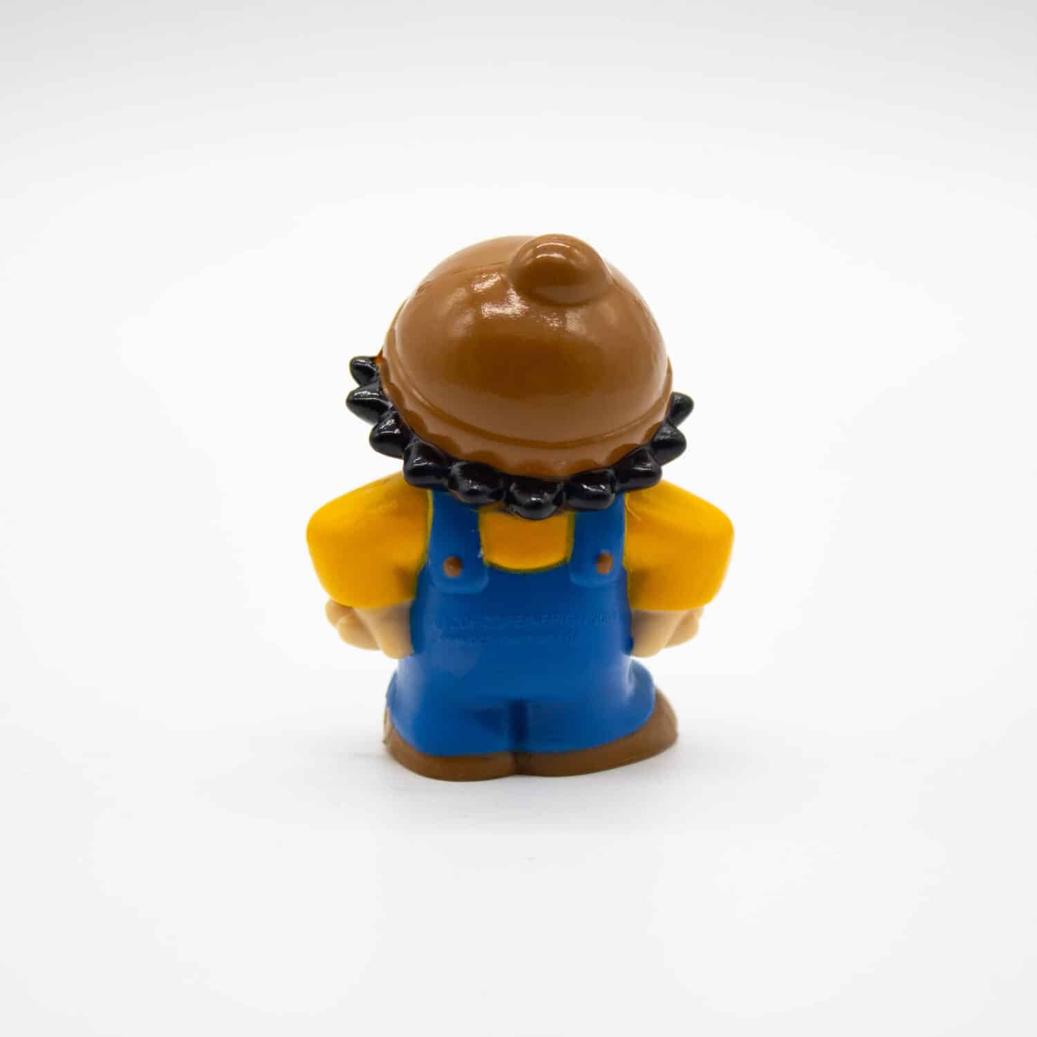 Plastikfigur eines Kindes in brauner Mütze, gelbem Oberteil und blauen Latzhosen | Rückseite